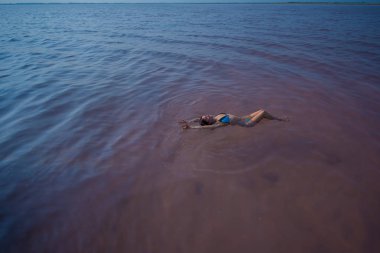 Mavi bikinili bir kadın tuz gölünde sırt larında yüzer. Doğanın mucizesi pembe göl. Tuz depozite ve çıkarma. Yoğun su yüzeye doğru itiyor. Maden suyunda tedavi prosedürleri.