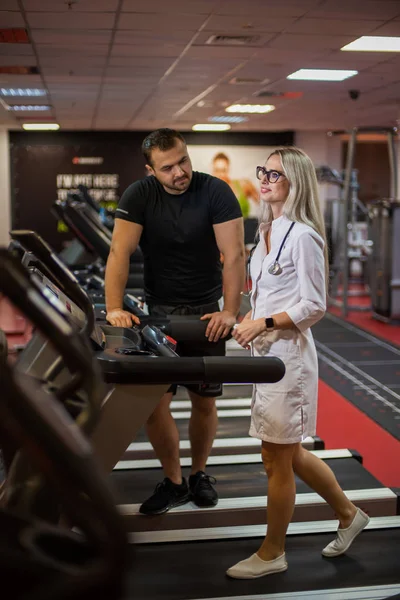 Sportowiec na siłowni kontroluje ćwiczenia bieżni pięknej lekarki w mundurze medycznym. Trener siłowni aprobuje smukłą sylwetkę pięknej fizjoterapeutki.. — Zdjęcie stockowe