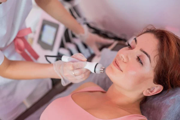 Jovem ruiva recebe uma massagem facial elétrica na luta contra o envelhecimento. Mulher de 30 anos recebe massagem facial ultrassônica elétrica contra rugas em um salão de beleza. Cosmetologia de hardware. — Fotografia de Stock