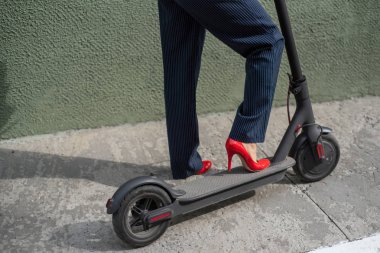 Kırmızı hight topuklu resmi giyim genç kadın elektrikli scooter üzerinde duruyor. Kadın bacaklarının yakın çekim. Pantolon kostümlü ve kırmızı ayakkabılı bir iş kadını elektrikli scooter ile şehrin etrafında dolaşıyor..