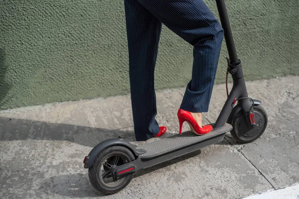 Giovane donna in abito formale sui tacchi alti rossi è in piedi su scooter elettrico. Primo piano delle gambe femminili. Una donna d'affari in giacca e cravatta e scarpe rosse si muove per la città su uno scooter elettrico . — Foto Stock