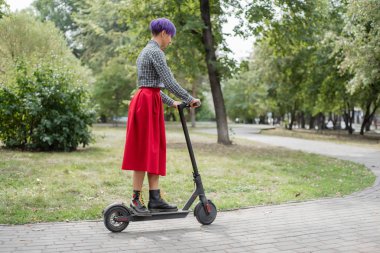 Mor saçlı genç bir kadın parkta elektrikli scooter kullanıyor. Ekose gömlek, uzun kırmızı etek ve papyon bir tıraş tapınak ile şık bir kız modern bir cihaz üzerinde şehir etrafında sürme. 