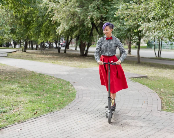 Eine junge Frau mit lila Haaren fährt mit einem Elektroroller in einem Park. ein stylisches Mädchen mit rasiertem Bügel im karierten Hemd, langem roten Rock und Fliege fährt auf einem modernen Gerät durch die Stadt. — Stockfoto