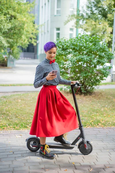 Μια νεαρή γυναίκα με μωβ μαλλιά καβαλάει ένα ηλεκτρικό σκούτερ στο πάρκο. Κομψό κορίτσι με ένα ξυρισμένο ναό σε ένα καρό πουκάμισο, μια μεγάλη κόκκινη φούστα και παπιγιόν γράφει ένα μήνυμα σε ένα smartphone. Hipster — Φωτογραφία Αρχείου