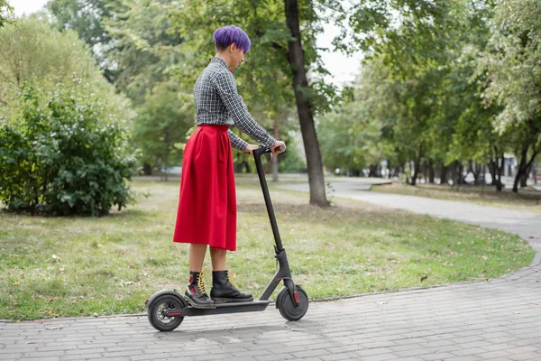 Mladá žena s purpurovými vlasy jede na elektrický skútr v parku. Stylová dívka s vyholenou chrámem v Kostkové košili, dlouhá rudá sukně a motýlek jezdí po městě na moderním zařízení. — Stock fotografie