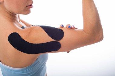 Beyaz arka planda Kineziotape bandı olan bir bayan omuz. Eklemlerde ve bağlarda spor sakatlıkları için alternatif tıp. Yaralı omzunda siyah bir tıbbi bantla genç bir sporcu. Kapat..