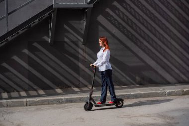 Beyaz gömlekli kızıl saçlı bir kız duvar boyunca elektrikli scooter kullanıyor. Pantolon kostümlü ve kırmızı topuklu ayakkabıgiyen bir iş kadını modern bir arabayla şehirde dolaşıyor. Scooter'da iş kadını.