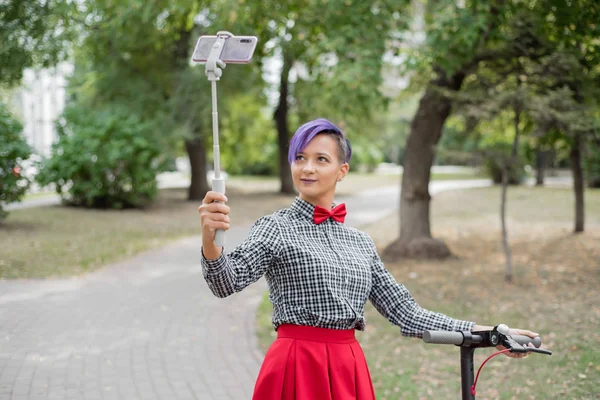 Eine junge Frau mit lila Haaren fährt mit einem Elektroroller in einem Park. Stilvolles Mädchen mit rasierter Schläfe im karierten Hemd, langem roten Rock und Fliege macht ein Foto mit einem Selfie-Stick. Hipster. — Stockfoto