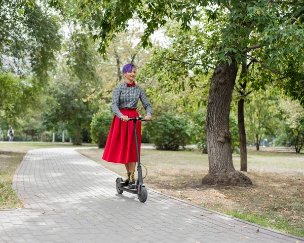 Молодая женщина с фиолетовыми волосами ездит на электрическом скутере в парке. Стильная девушка в клетчатой рубашке, длинной красной юбке и галстуке-бабочке разъезжает по городу на современном устройстве. Хипстер . — стоковое фото