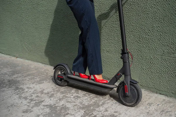 Jonge vrouw in formele slijtage op rode hight hakken staat op elektrische scooter. Close-up van vrouwelijke benen. Een zakenvrouw in een broekpak en rode schoenen beweegt zich rond de stad op een elektrische scooter. — Stockfoto