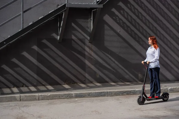Una ragazza dai capelli rossi con una camicia bianca guida uno scooter elettrico lungo il muro. Una donna d'affari in giacca e cravatta e tacchi rossi gira per la città in un'auto moderna. Donna d'affari su uno scooter . — Foto Stock