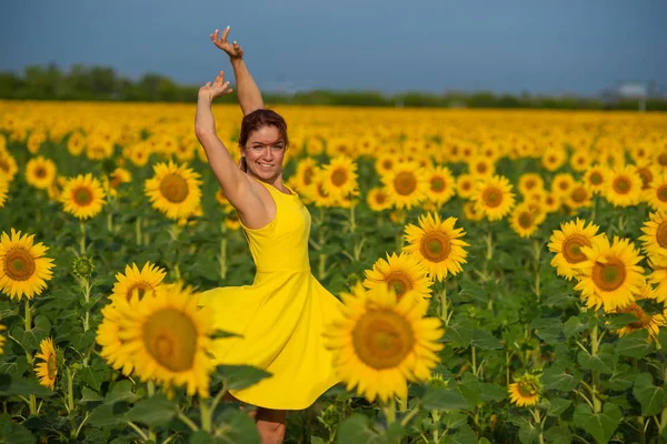 Rothaarige Frau in einem gelben Kleid, die mit erhobenen Händen in einem Sonnenblumenfeld tanzt. schöne Mädchen im Rock Sonne genießt einen wolkenlosen Tag auf dem Land. rosa Haarsträhnen. — Stockfoto