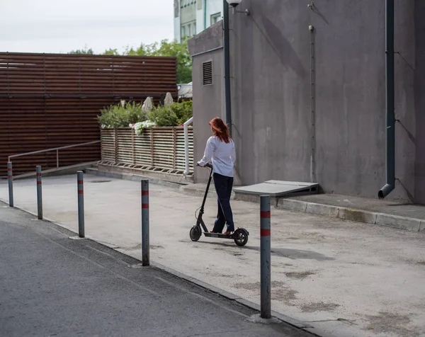 Beyaz gömlekli kızıl saçlı bir kız duvar boyunca elektrikli scooter kullanıyor. Pantolon kostümlü ve kırmızı topuklu ayakkabıgiyen bir iş kadını modern bir arabayla şehirde dolaşıyor. Scooter'da iş kadını. — Stok fotoğraf