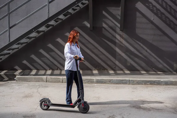 Una chica pelirroja con camisa blanca conduce un scooter eléctrico a lo largo de la pared. Una mujer de negocios con un traje de pantalón y tacones altos rojos pasea por la ciudad en un coche moderno. Mujer de negocios en un scooter . — Foto de Stock