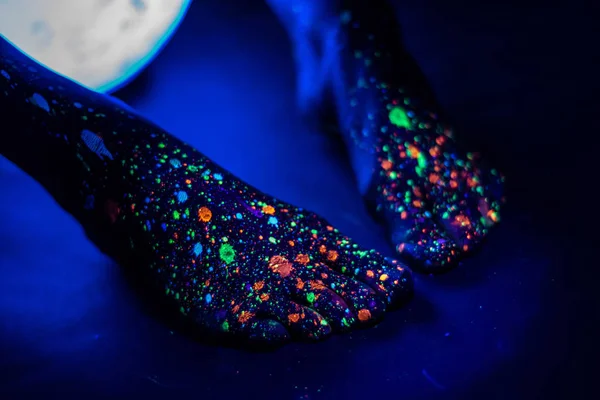 Piedi femminili scalzi in uno spruzzo di vernice incandescente al buio. Dita in polvere fluorescente. Body art incandescente nella luce ultravioletta, come un cielo notturno blu con stelle. Da vicino. . — Foto Stock