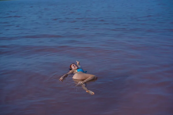 Μια γυναίκα με μπλε μπικίνι κολυμπάει στην πλάτη της σε μια αλμυρή λίμνη. Το θαύμα της ροζ λίμνης της φύσης. Κατάθεση και εξαγωγή αλατιού. Πυκνό νερό που πιέζει στην επιφάνεια. Θεραπευτικές διαδικασίες σε μεταλλικό νερό. — Φωτογραφία Αρχείου