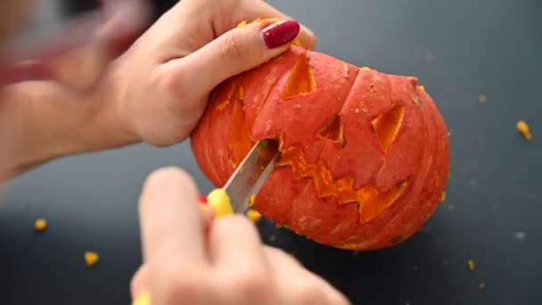 Uma mulher esculpe um grimace maligno em uma abóbora para o Halloween com uma faca de papel. Close up de mãos femininas fazendo jack-o-lanterna em uma mesa preta na véspera de todos os santos . — Vídeo de Stock