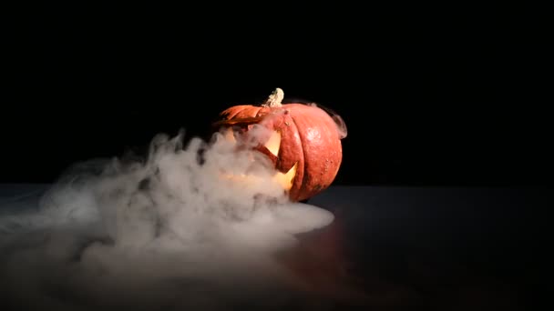 Halloween, oranje pompoen met een eng licht gezicht op een donkere achtergrond. Dikke grijze rook komt naar buiten en verspreidt zich over de zwarte tafel. Een close-up van een zaklamp aan de vooravond van alle heiligen — Stockvideo