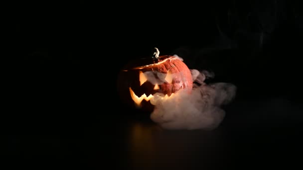 Halloween, abóbora laranja com um rosto luminoso assustador em um fundo escuro. O fumo cinzento espesso sai e espalha-se pela mesa preta. Um close-up de uma lanterna na véspera de todos os santos — Vídeo de Stock