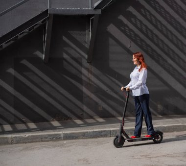 Beyaz gömlekli kızıl saçlı bir kız duvar boyunca elektrikli scooter kullanıyor. Pantolon kostümlü ve kırmızı topuklu ayakkabıgiyen bir iş kadını modern bir arabayla şehirde dolaşıyor. Scooter'da iş kadını.
