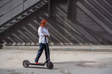 Beyaz gömlekli kızıl saçlı bir kız duvar boyunca elektrikli scooter kullanıyor. Pantolon kostümlü ve kırmızı topuklu ayakkabıgiyen bir iş kadını modern bir arabayla şehirde dolaşıyor. Scooter'da iş kadını.