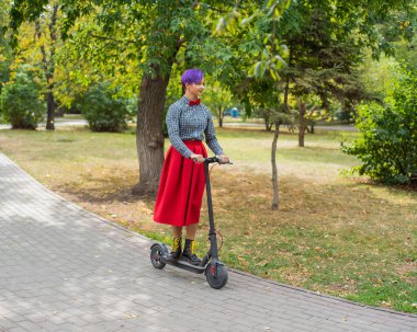 Mor saçlı genç bir kadın parkta elektrikli scooter kullanıyor. Ekose gömlek, uzun kırmızı etek ve papyon şık kız modern bir cihaz üzerinde şehir etrafında sürme. Hipster.