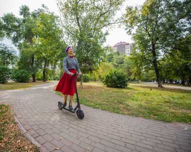 Mor saçlı genç bir kadın parkta elektrikli scooter kullanıyor. Ekose gömlek, uzun kırmızı etek ve papyon bir tıraş tapınak ile şık bir kız modern bir cihaz üzerinde şehir etrafında sürme. 