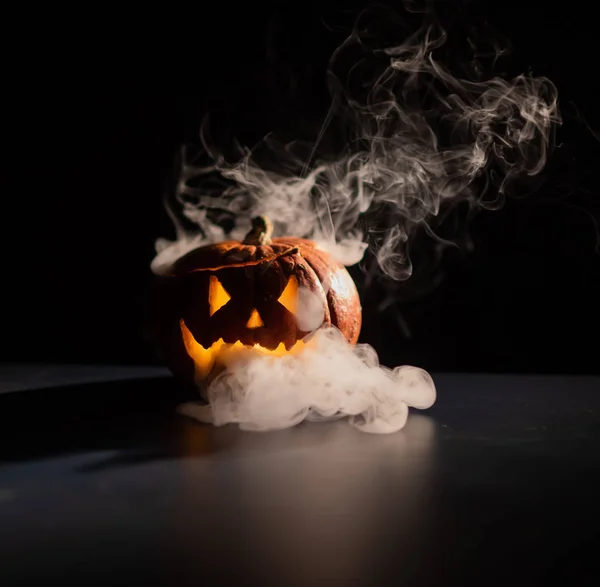 Halloween, oranje pompoen met een eng licht gezicht op een donkere achtergrond. Dikke grijze rook komt naar buiten en verspreidt zich over de zwarte tafel. Een close-up van een zaklamp. Versierde kaars stand. — Stockfoto