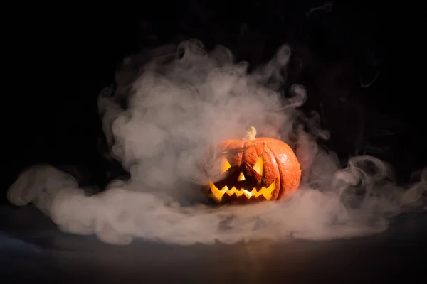 Halloween, orange pumpkin with a scary luminous face on a dark background. Thick gray smoke comes out and spreads across the black table. A close-up of a flashlight on the eve of all the saints — Stock Photo, Image