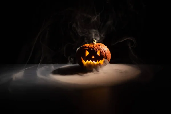 Halloween, orange pumpkin with a scary luminous face on a dark background. Thick gray smoke comes out and spreads across the black table. A close-up of a flashlight on the eve of all the saints — Stock Photo, Image