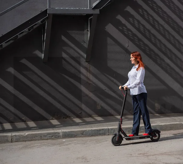 A red-haired girl in a white shirt drives an electric scooter along the wall. A business woman in a trouser suit and red high heels rides around the city in a modern car. Business woman on a scooter.