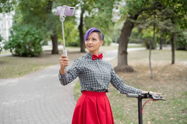 Eine junge Frau mit lila Haaren fährt mit einem Elektroroller in einem Park. Stilvolles Mädchen mit rasierter Schläfe im karierten Hemd, langem roten Rock und Fliege macht ein Foto mit einem Selfie-Stick. Hipster. — Stockfoto