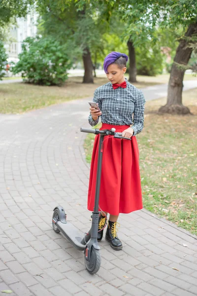 A fiatal nő, lila hajú lovaglás elektromos robogó a parkban. Stílusos lány egy borotvált templom egy kockás ing, egy hosszú vörös szoknya és csokornyakkendő írja egy üzenetet a smartphone. Csípő — Stock Fotó