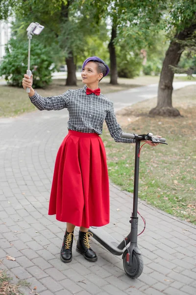 Eine junge Frau mit lila Haaren fährt mit einem Elektroroller in einem Park. Stilvolles Mädchen mit rasierter Schläfe im karierten Hemd, langem roten Rock und Fliege macht ein Foto mit einem Selfie-Stick. Hipster. — Stockfoto