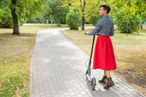 Eine junge Frau mit lila Haaren fährt mit einem Elektroroller in einem Park. ein stylisches Mädchen mit rasiertem Bügel im karierten Hemd, langem roten Rock und Fliege fährt auf einem modernen Gerät durch die Stadt. — Stockfoto