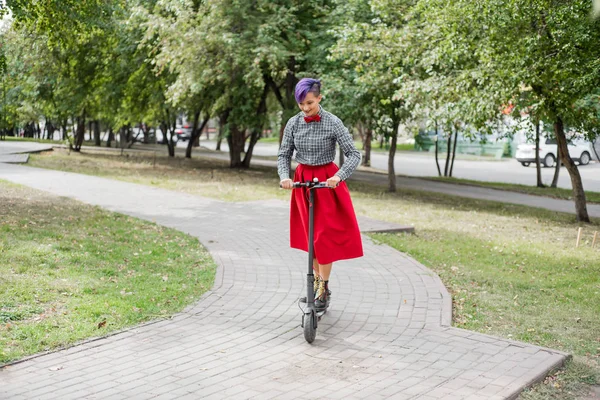 Uma jovem com cabelo roxo monta uma scooter elétrica em um parque. Uma menina elegante com um templo raspado em uma camisa xadrez, uma saia vermelha longa e um laço está andando ao redor da cidade em um dispositivo moderno . — Fotografia de Stock