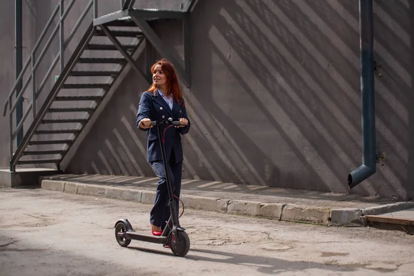 Ceket ve pantolon giyen kızıl saçlı bir kız duvar boyunca elektrikli scooter kullanıyor. Bir pantsuit ve kırmızı yüksek topuklu bir iş kadını modern bir araba ile şehir etrafında sürmek. Ofis çalışanı. Kıyafet kodu — Stok fotoğraf