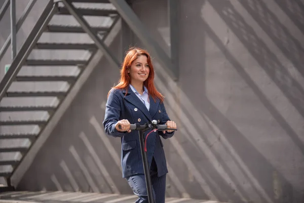 A red-haired girl in a jacket and trousers drives an electric scooter along the wall. A business woman in a pantsuit and red high heels rides around the city in a modern car. Office worker. Dress code