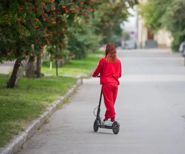 Chica pelirroja con un chándal rojo conduce un scooter eléctrico. Una joven con ropa de gran tamaño pasea por la ciudad y escucha música usando auriculares inalámbricos. Vista desde atrás . —  Fotos de Stock