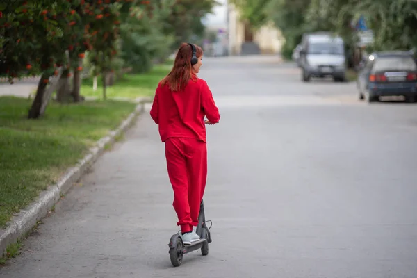 Chica pelirroja con un chándal rojo conduce un scooter eléctrico. Una joven con ropa de gran tamaño pasea por la ciudad y escucha música usando auriculares inalámbricos. Vista desde atrás . — Foto de Stock