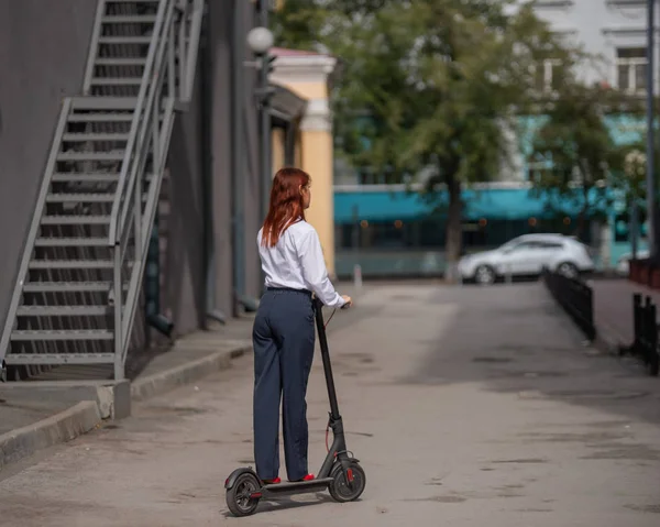 Rudowłosy dziewczyna w białej koszulce napędza skuter elektryczny wzdłuż ściany. Kobieta biznesu w garniturze spodni i czerwone wysokie obcasy jeździ po mieście w nowoczesnym samochodzie. kod stroju w biurze. — Zdjęcie stockowe