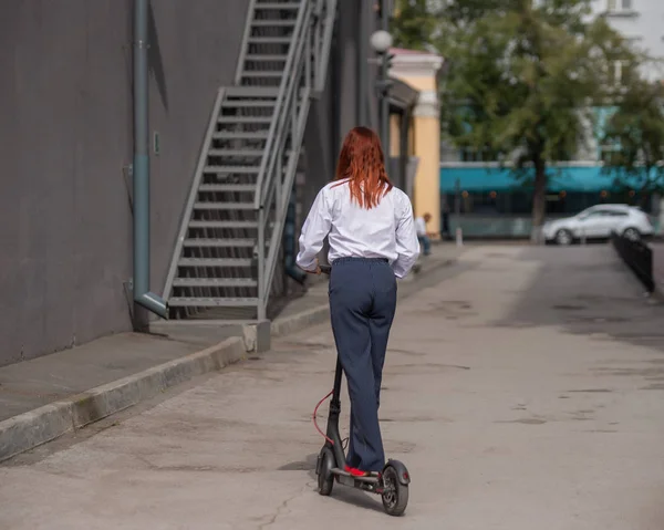 Рыжая девушка в белой рубашке водит электрический скутер вдоль стены. Деловая женщина в брючном костюме и на красных каблуках разъезжает по городу на современной машине. дресс-код в офисе . — стоковое фото