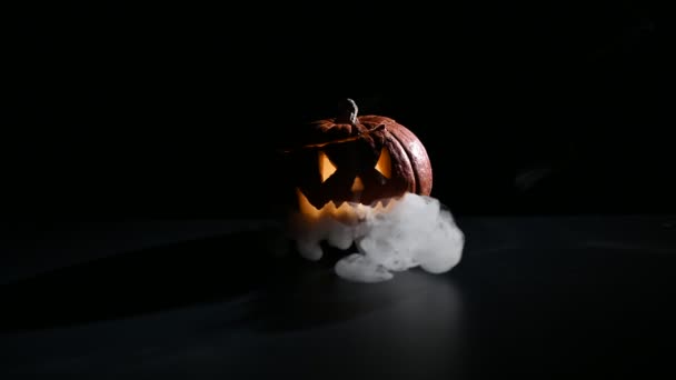 Halloween, abóbora laranja com um rosto luminoso assustador em um fundo escuro. O fumo cinzento espesso sai e espalha-se pela mesa preta. Um close-up de uma lanterna na véspera de todos os santos — Vídeo de Stock