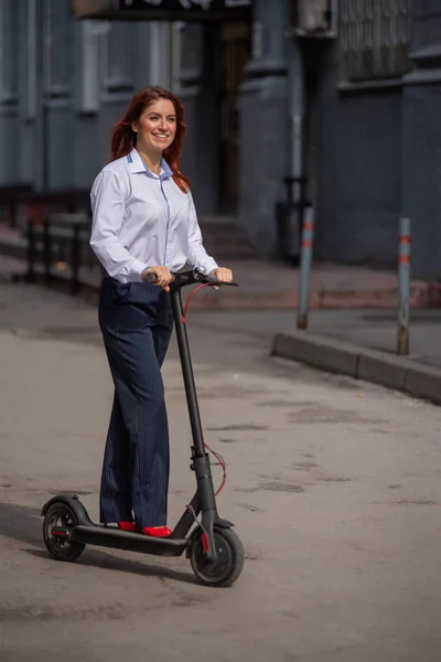 Una chica pelirroja con camisa blanca conduce un scooter eléctrico a lo largo de la pared. Una mujer de negocios con un traje de pantalón y tacones altos rojos pasea por la ciudad en un coche moderno. código de vestimenta en la oficina . — Foto de Stock