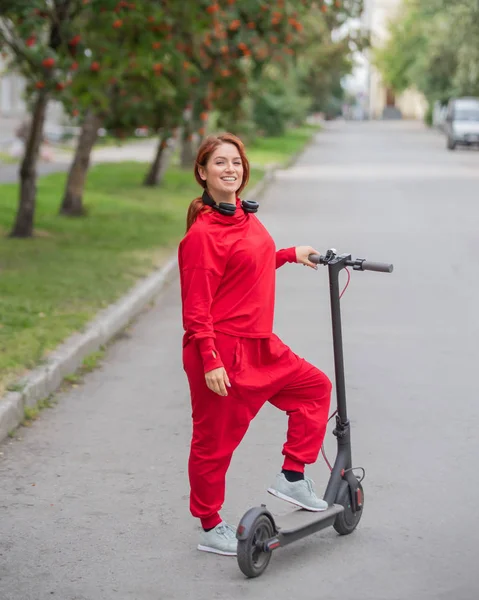 Chica pelirroja con un chándal rojo conduce un scooter eléctrico. Una joven en ropa de gran tamaño pasea por la ciudad en transporte moderno y escucha música usando auriculares inalámbricos . — Foto de Stock