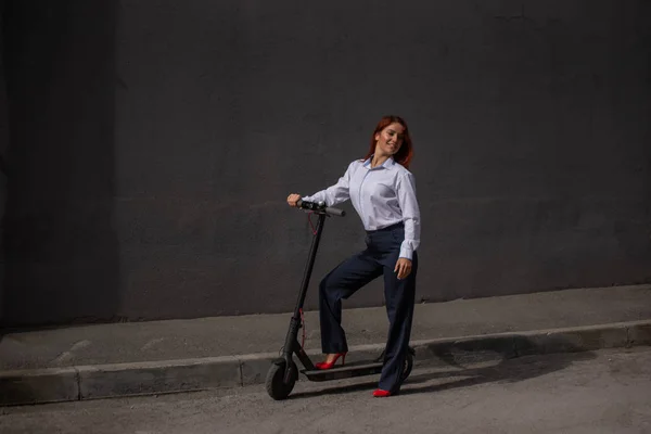 Una ragazza dai capelli rossi con una camicia bianca guida uno scooter elettrico lungo il muro. Una donna d'affari in giacca e cravatta e tacchi rossi gira per la città in un'auto moderna. dress code in ufficio . — Foto Stock