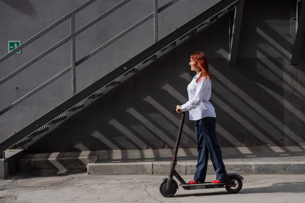 Una chica pelirroja con camisa blanca conduce un scooter eléctrico a lo largo de la pared. Una mujer de negocios con un traje de pantalón y tacones altos rojos pasea por la ciudad en un coche moderno. Mujer de negocios en un scooter . — Foto de Stock