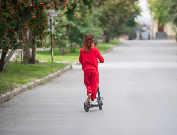 Rudowłosy dziewczyna w czerwonym dres napędza skuter elektryczny. Młoda kobieta w przewymiarowany ubrania jeździ po mieście i słucha muzyki za pomocą słuchawek bezprzewodowych. Widok z tyłu. — Zdjęcie stockowe