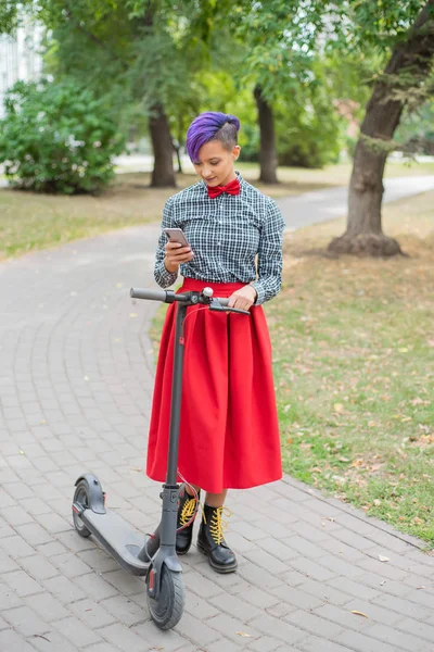 Eine junge Frau mit lila Haaren fährt im Park auf einem Elektroroller. Stilvolles Mädchen mit rasierter Schläfe im karierten Hemd, langem roten Rock und Fliege schreibt eine Nachricht auf ein Smartphone. Hipster — Stockfoto