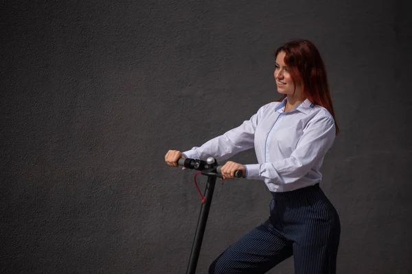 A red-haired girl in a white shirt drives an electric scooter along the wall. A business woman in a trouser suit and red high heels rides around the city in a modern car. dress code in the office.